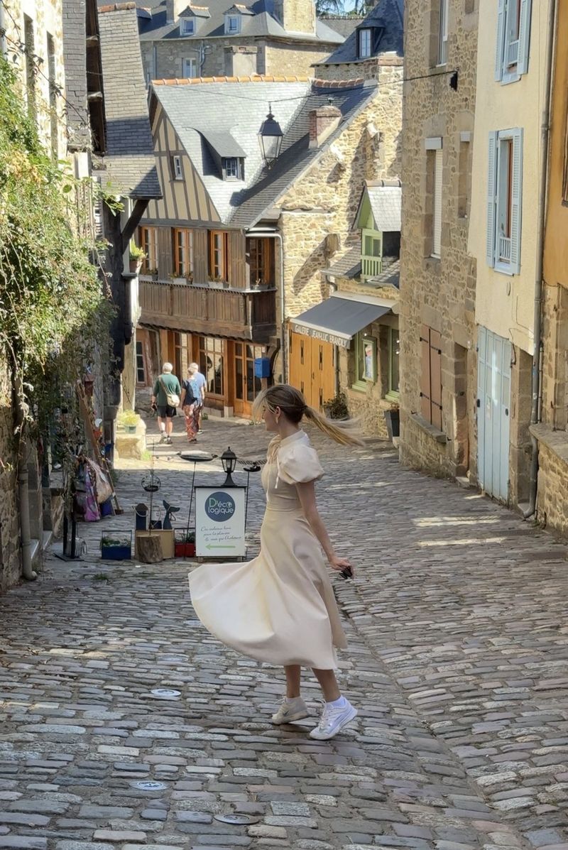 A woman in a white dress twirling on a steep cobbled street lined with half-timbered and stone houses in Dinan