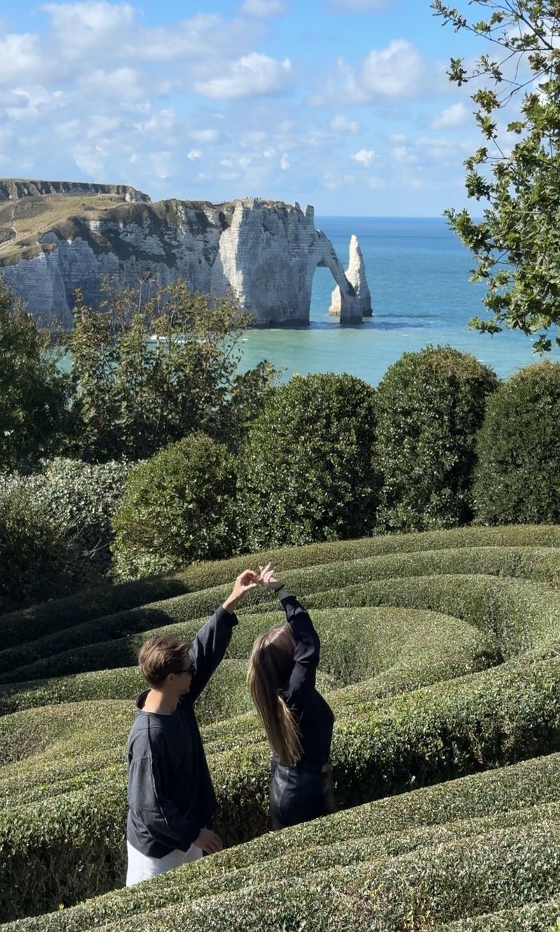 A couple making a heart with their hands among wave-shaped clipped hedges in Les Jardins d’Étretat, with the white cliffs and the arch behind