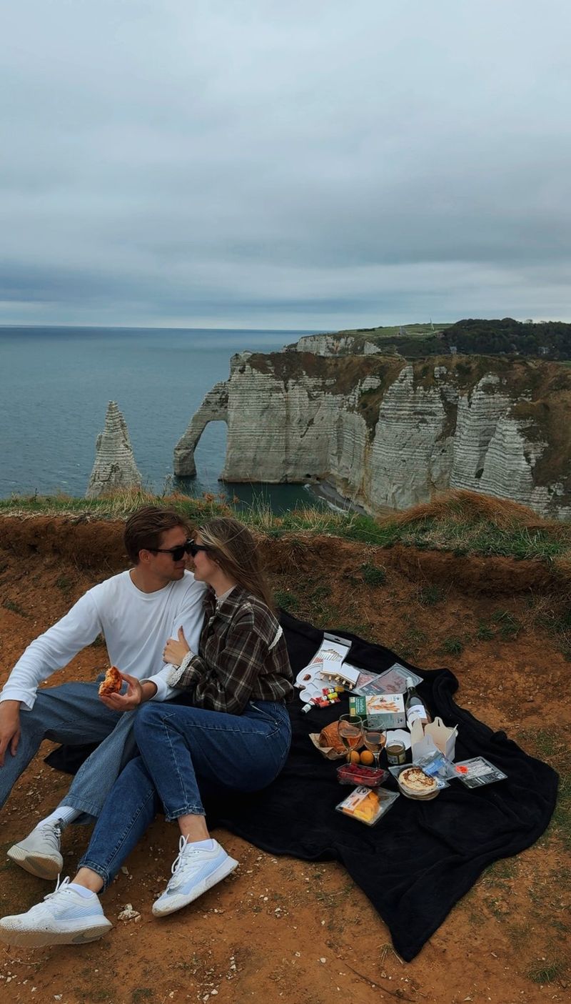 A couple kissing on a picnic blanket on the clifftop at Étretat, with the arch and the Needle behind them
