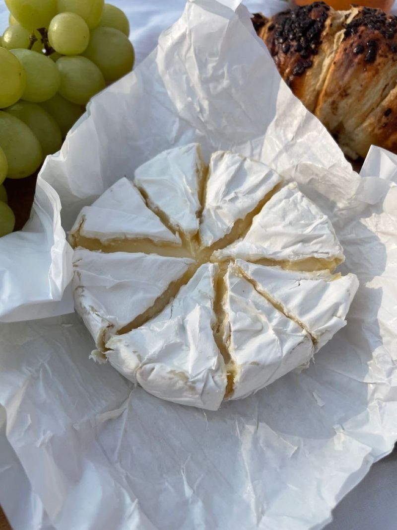 A round Norman cheese cut into wedges on white paper, with green grapes beside it
