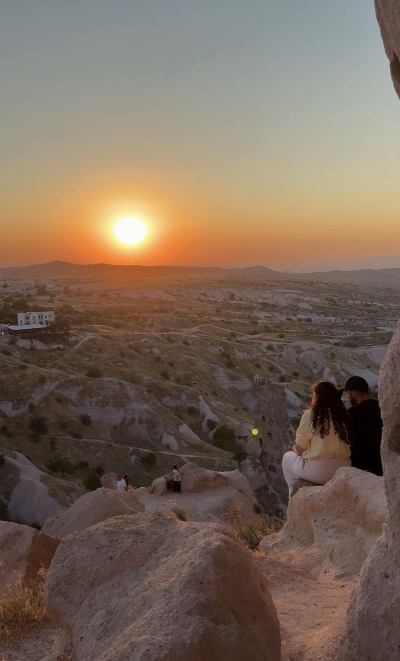 A couple sitting on a rock ledge above the valleys of Uçhisar, watching an orange sun set over the hills