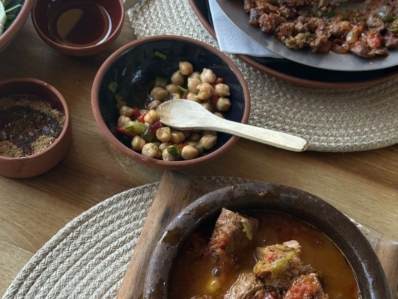 A wooden table seen from above: a bowl of chickpea salad with a wooden spoon, small dishes of spices and a clay pot of meat stew