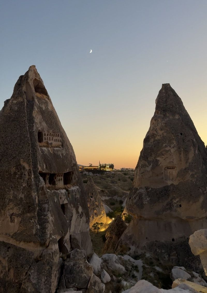 Two tall fairy chimneys with carved windows framing a valley at dusk, a thin crescent moon in the sky above
